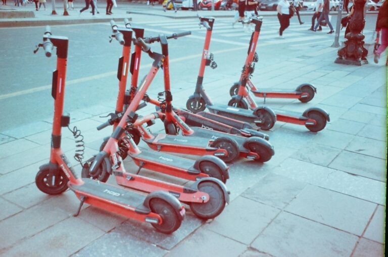 Red electric scooters lined up on an urban sidewalk with pedestrians in the background.